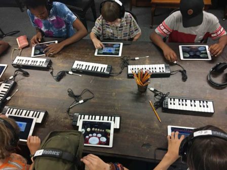 A group of six students of different ethnicities play music with tiny keyboards and iPads.