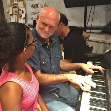 An older man sits at a piano, teaching piano to a young girl.